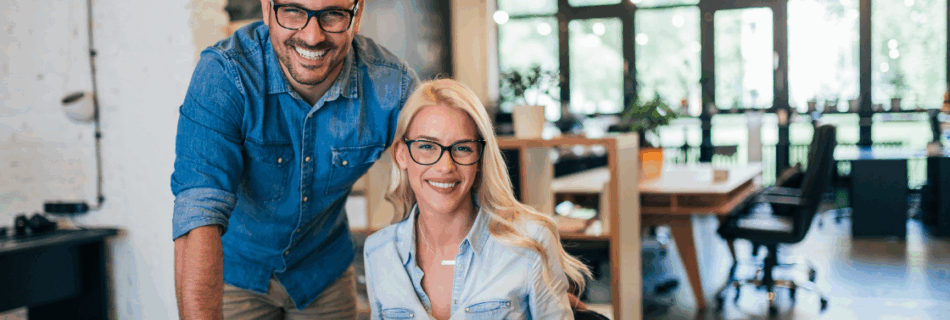 woman sitting at a desk with a laptop and a man standing next to her both smiling Why Proactive Business Owners Never Scramble at Tax Season (And How You Can Join Them)