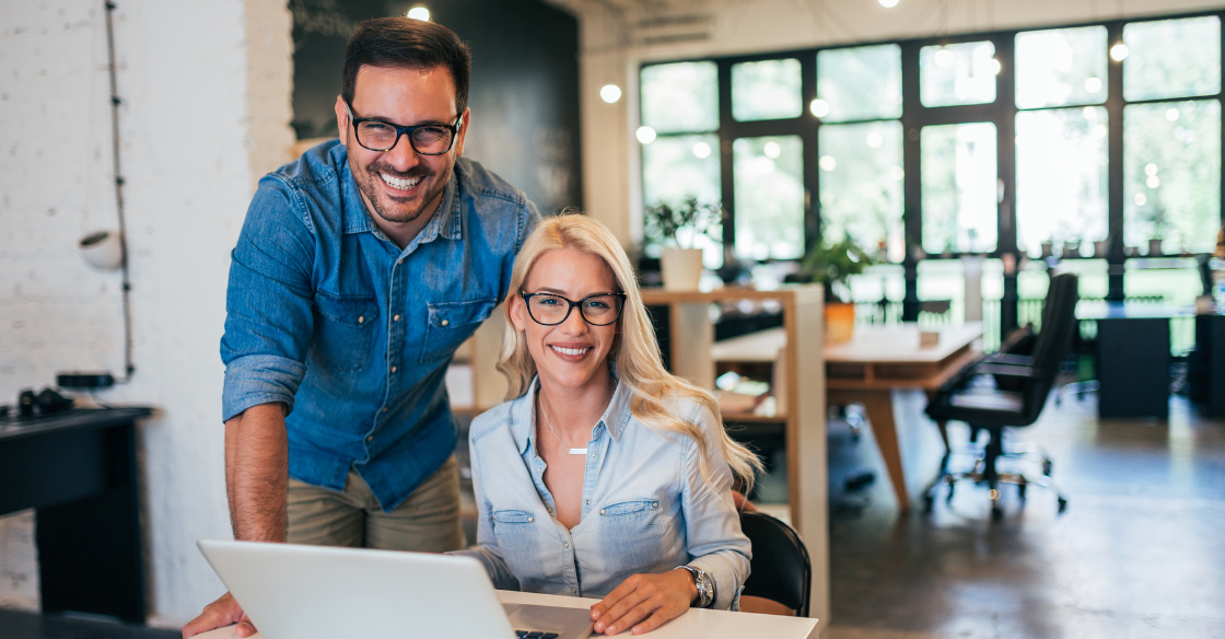 woman sitting at a desk with a laptop and a man standing next to her both smiling Why Proactive Business Owners Never Scramble at Tax Season (And How You Can Join Them)