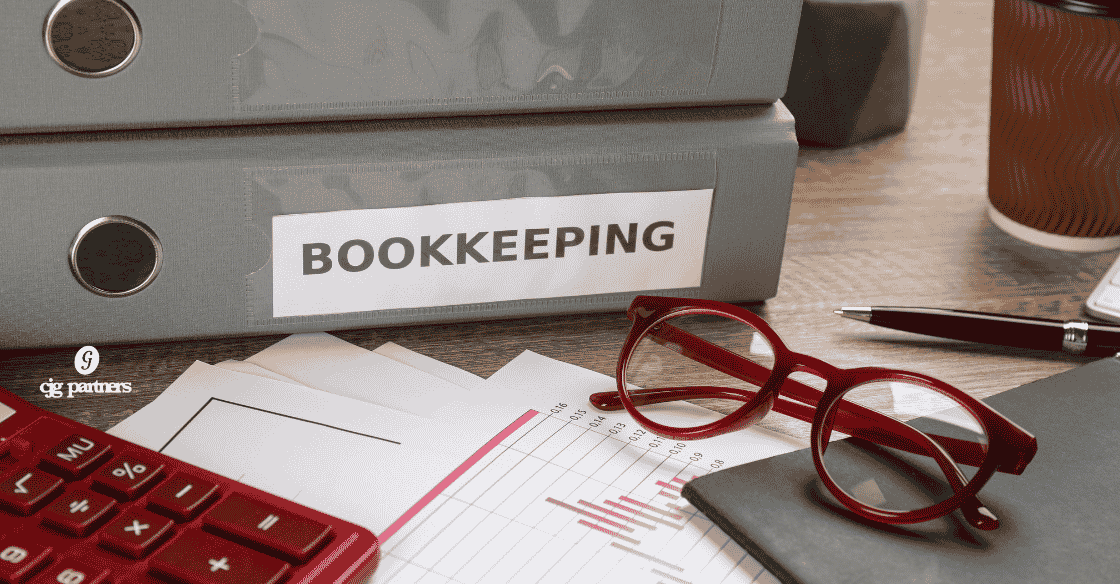 binders with bookkeeping on the spine and papers, glasses, and a calculator on a table The Unexpected Value in Consistent Bookkeeping cjg partners